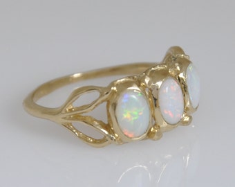 May include: A gold ring featuring three oval-shaped opals set in a row. The band has a decorative, open-work design that branches out toward the settings. Each opal displays a milky white base color with iridescent flashes of green, orange, and red.