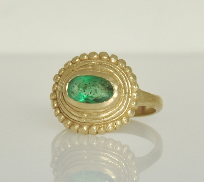 May include: A gold ring with a green emerald gemstone set in a textured oval bezel. The ring has a simple band.