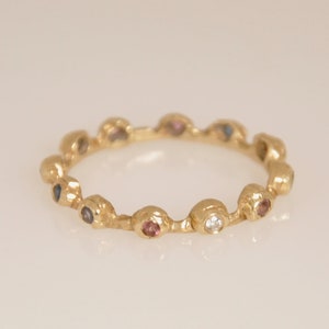 May include: A gold band ring with a row of colorful gemstones. The ring features a variety of small, round stones in shades of blue, pink, and white.