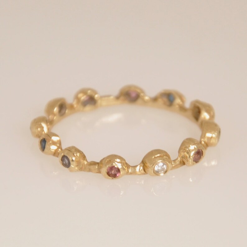 May include: A gold band ring with a row of colorful gemstones. The ring features a variety of small, round stones in shades of blue, pink, and white.