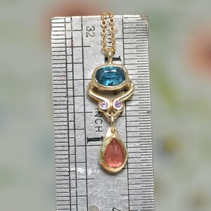 May include: A gold pendant necklace with a blue and green gemstone, two smaller pink gemstones, and a dangling red gemstone. The necklace is measured against a ruler.