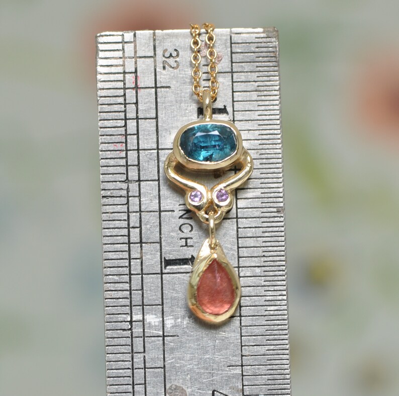 May include: A gold pendant necklace with a blue and green gemstone, two smaller pink gemstones, and a dangling red gemstone. The necklace is measured against a ruler.