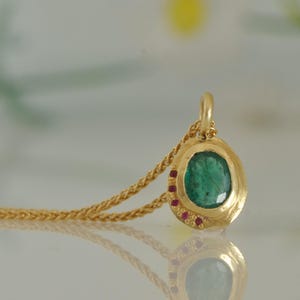 May include: Gold pendant necklace featuring a large, oval-shaped emerald gemstone. The pendant is surrounded by a gold bezel and accented with small red gemstones. The necklace chain is also gold.