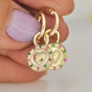 May include: Gold hoop earrings with a heart charm. The charm is surrounded by a circle of pink and green gemstones.