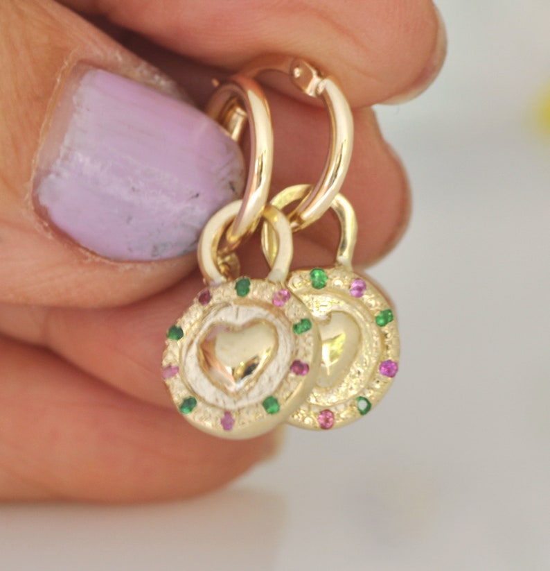 May include: Gold hoop earrings with a heart charm. The charm is surrounded by a circle of pink and green gemstones.