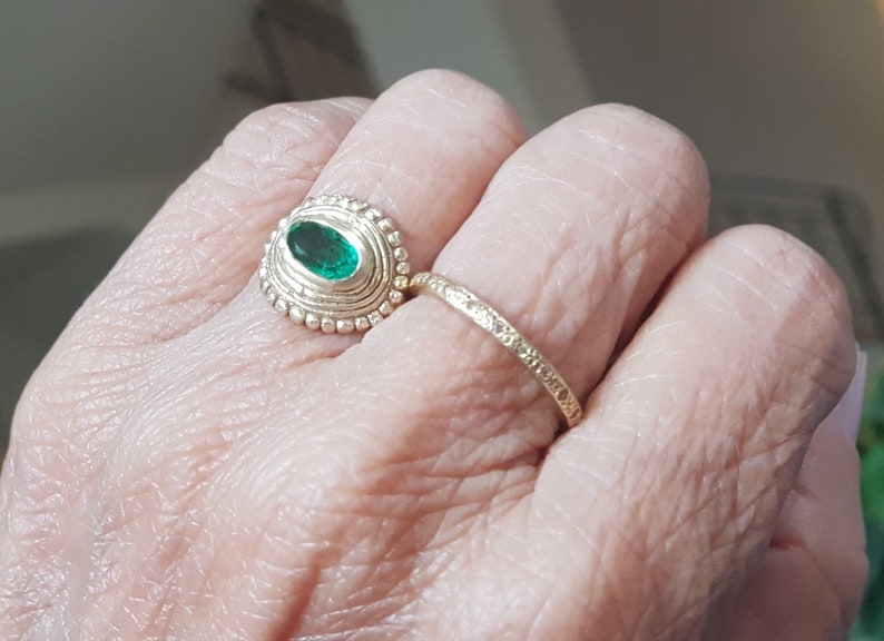 May include: A gold ring with a green oval gemstone set in a beaded bezel. A second, thinner gold band is worn on the same finger.