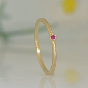 May include: A delicate gold ring with a small, round, bright pink gemstone. The ring has a thin band and a simple, elegant design, perfect for everyday wear or special occasions. The gemstone is set in a minimalist style.