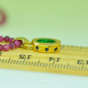 Pu&ograve; includere: Primo piano di un ciondolo dorato con una grande gemma verde al centro e piccole gemme blu. Il ciondolo &egrave; attaccato a una collana di perline con piccole gemme rosa. Il ciondolo &egrave; su un righello di legno.