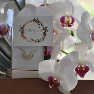 Puede incluir: Un collar de oro con un delicado colgante que deletrea la palabra "Trust" en una fuente cursiva. El collar se muestra en una caja blanca con un dise&ntilde;o floral y el texto "Mullen Russworm Jewelry". La caja est&aacute; rodeada de orqu&iacute;deas blancas con centros rosados.