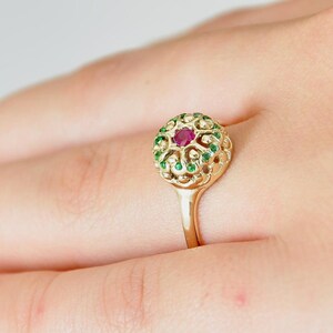 May include: A gold ring with a red ruby stone surrounded by green gemstones. The ring has a delicate filigree design.