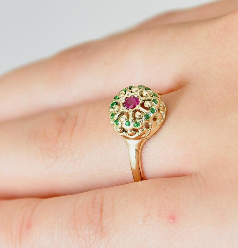May include: A gold ring with a red ruby stone surrounded by green gemstones. The ring has a delicate filigree design.