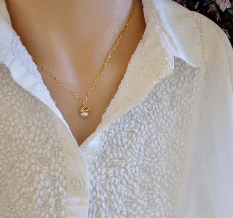 May include: A delicate gold chain necklace with a small pearl pendant. The necklace is worn by a person wearing a white blouse with an embroidered pattern.
