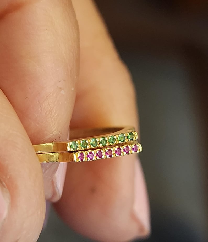 May include: Two gold rings with a row of small green and pink gemstones. The rings are stacked on top of each other.