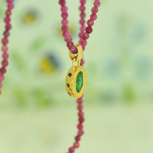 Pu&ograve; includere: Un pendente dorato con una gemma verde al centro e piccoli accenti blu scuro, sospeso da una collana di piccole perline rosa rotonde. Il pendente &egrave; di forma ovale e ha una cornice dorata strutturata. Lo sfondo &egrave; verde tenue con motivi floreali sfocati.