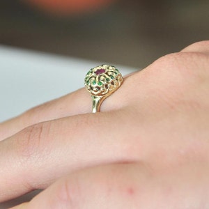 May include: A gold ring with a red and green gemstone setting. The ring is on a person's finger.