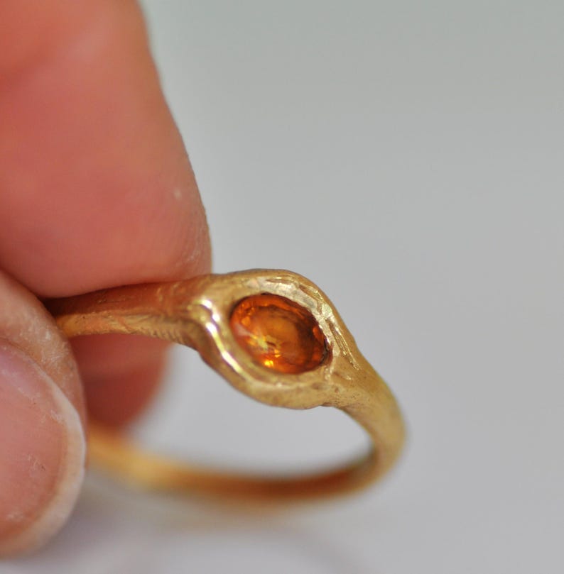 May include: A gold ring with an oval, orange gemstone. The ring has a textured band and a bezel setting that holds the stone. The ring is held by a person's fingers.