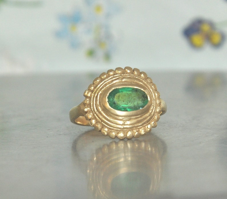 May include: A gold ring with a green emerald gemstone set in a textured, circular bezel. The ring has a simple band.