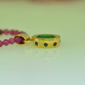 Pu&ograve; includere: Un pendente in oro con una pietra centrale rotonda color smeraldo e piccole pietre blu. Il pendente &egrave; attaccato a un filo di piccole perline rosa. Il pendente ha una superficie strutturata.