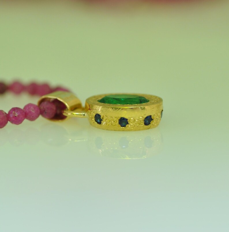 Pu&ograve; includere: Un pendente in oro con una pietra centrale rotonda color smeraldo e piccole pietre blu. Il pendente &egrave; attaccato a un filo di piccole perline rosa. Il pendente ha una superficie strutturata.