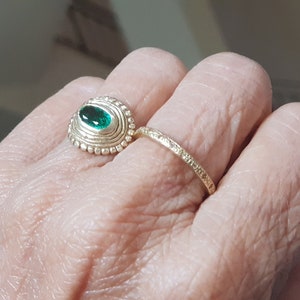 May include: A gold ring with a green gemstone set in a textured oval bezel. The ring is worn on a finger with a second, thinner gold band.