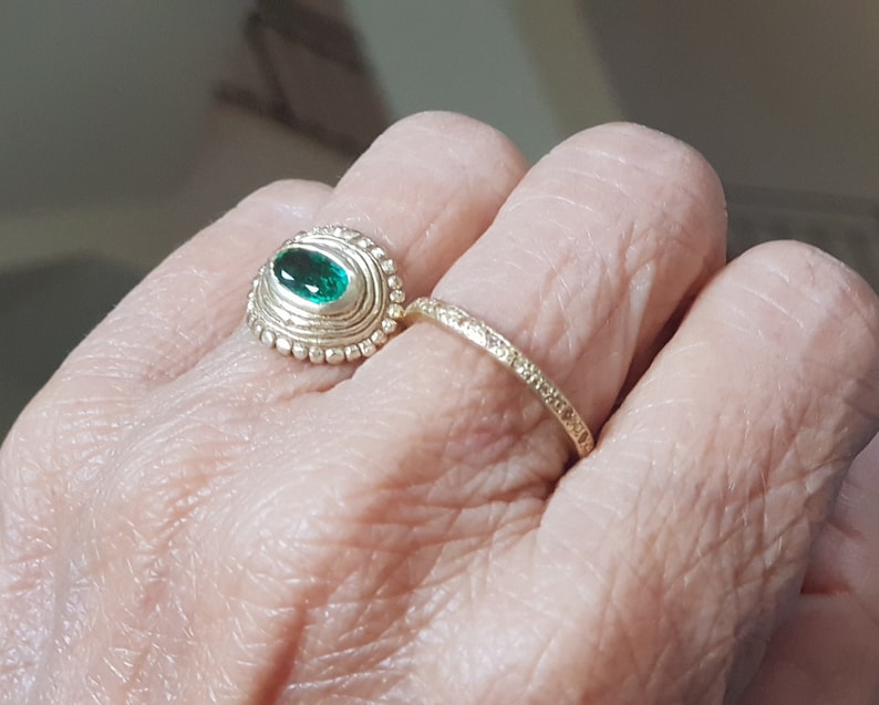 May include: A gold ring with a green gemstone set in a textured oval bezel. The ring is worn on a finger with a second, thinner gold band.