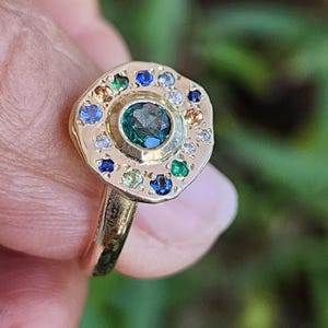 K&ouml;nnte beinhalten: Ein goldener Ring mit einem gr&uuml;nen Edelstein, der von einem Heiligenschein aus kleineren bunten Edelsteinen umgeben ist.