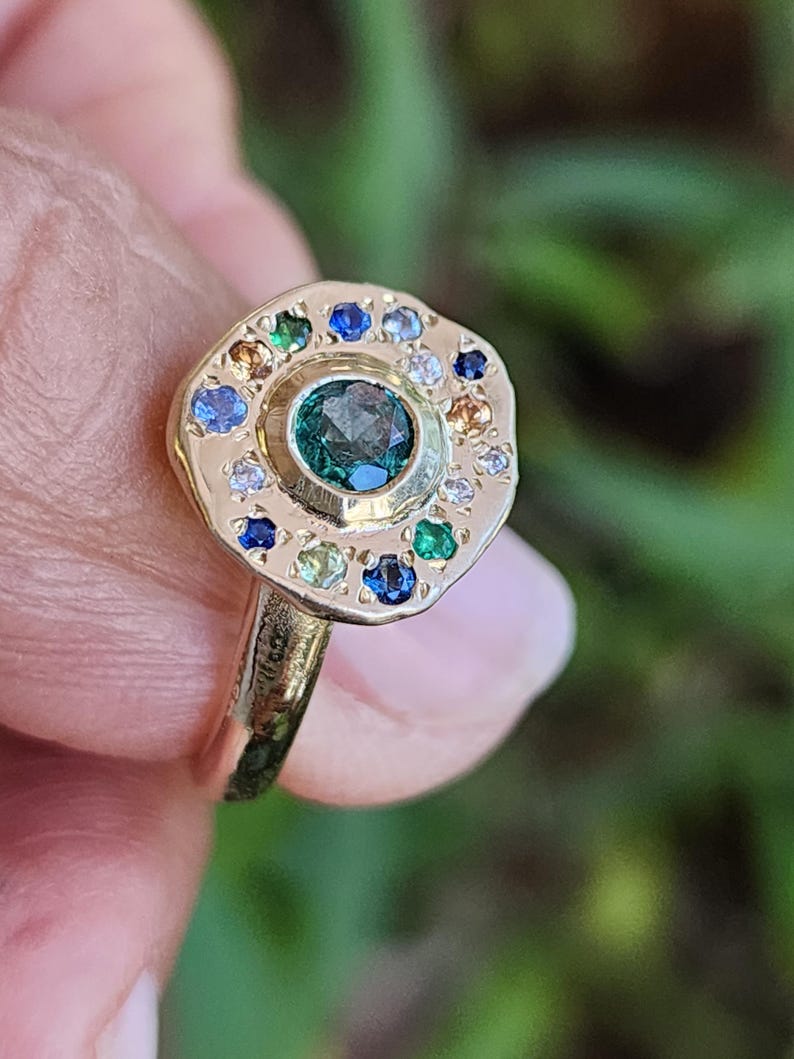 K&ouml;nnte beinhalten: Ein goldener Ring mit einem gr&uuml;nen Edelstein, der von einem Heiligenschein aus kleineren bunten Edelsteinen umgeben ist.