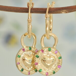 May include: Gold hoop earrings with a heart-shaped charm. The charm is surrounded by a circle of green and pink gemstones.