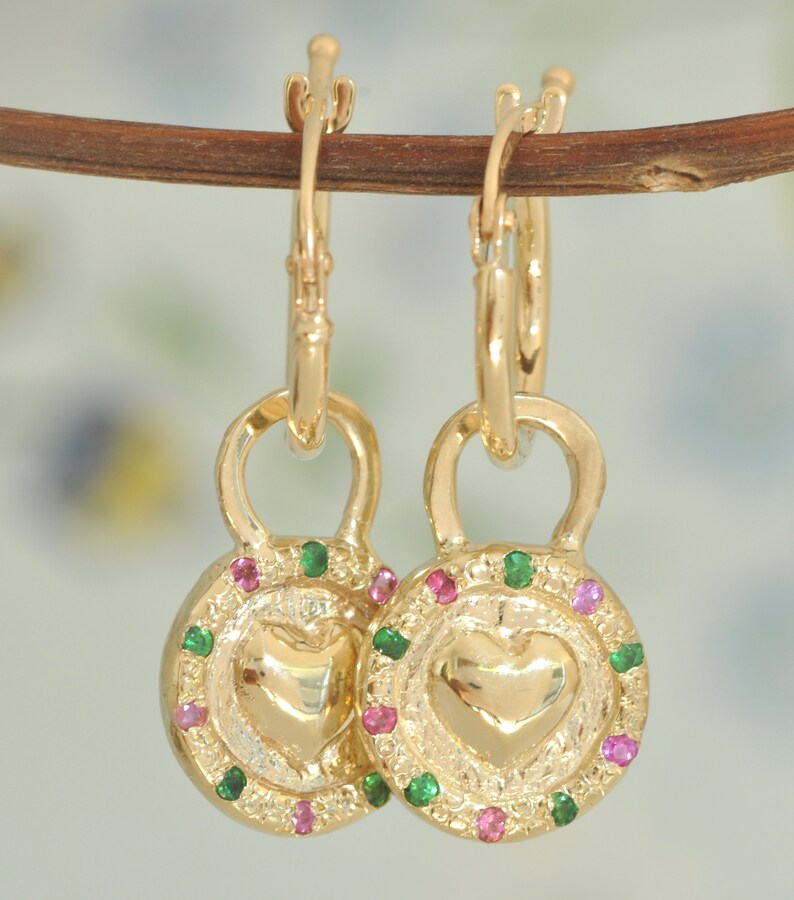 May include: Gold hoop earrings with a heart-shaped charm. The charm is surrounded by a circle of green and pink gemstones.