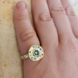 K&ouml;nnte beinhalten: Ein goldener Ring mit einer runden, mehrfarbigen Edelsteinbesetzung. Der Ring hat einen gro&szlig;en, gr&uuml;nen Edelstein in der Mitte, umgeben von kleineren, blauen, gelben und wei&szlig;en Edelsteinen.