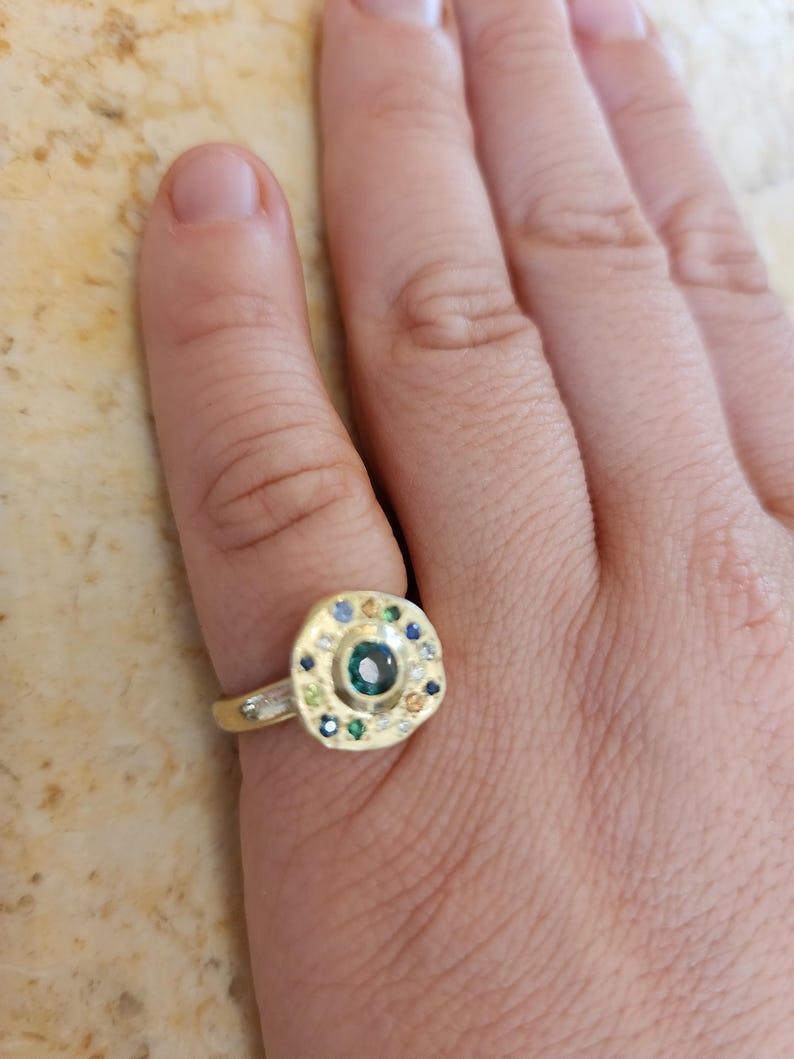 K&ouml;nnte beinhalten: Ein goldener Ring mit einer runden, mehrfarbigen Edelsteinbesetzung. Der Ring hat einen gro&szlig;en, gr&uuml;nen Edelstein in der Mitte, umgeben von kleineren, blauen, gelben und wei&szlig;en Edelsteinen.