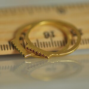 May include: Two gold rings with a row of small red gemstones on each. The rings are stacked on top of each other and are resting on a wooden ruler.