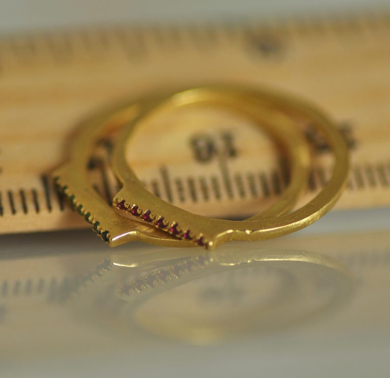 May include: Two gold rings with a row of small red gemstones on each. The rings are stacked on top of each other and are resting on a wooden ruler.