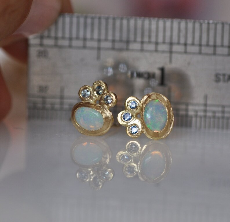 Op de afbeelding: Een paar gouden oorbellen met ovale opalen en kleine blauwe edelstenen. De oorbellen zijn geplaatst tegen een witte achtergrond.