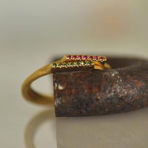 May include: A gold ring with a thin band and a double row of small gemstones. The top row of gemstones is pink, and the bottom row is green.
