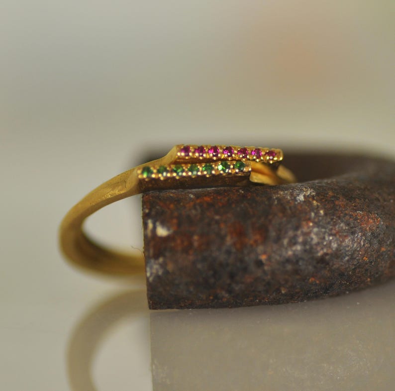 May include: A gold ring with a thin band and a double row of small gemstones. The top row of gemstones is pink, and the bottom row is green.