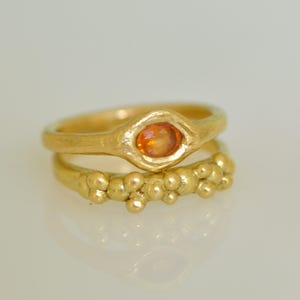 May include: Two gold rings. The top ring has an oval-shaped orange gemstone. The bottom ring has a textured band with small, round gold accents. Both rings are set against a white background.