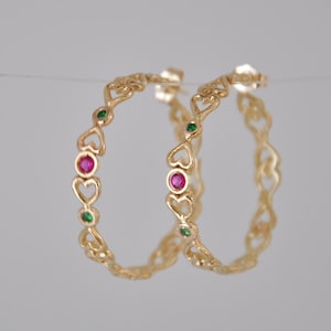 May include: Gold hoop earrings with a heart-shaped design and alternating pink and green gemstones.
