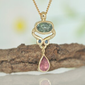 May include: A gold pendant necklace with a teardrop-shaped pink gemstone, a green oval gemstone, and two small blue gemstones. The pendant is hanging from a gold chain.