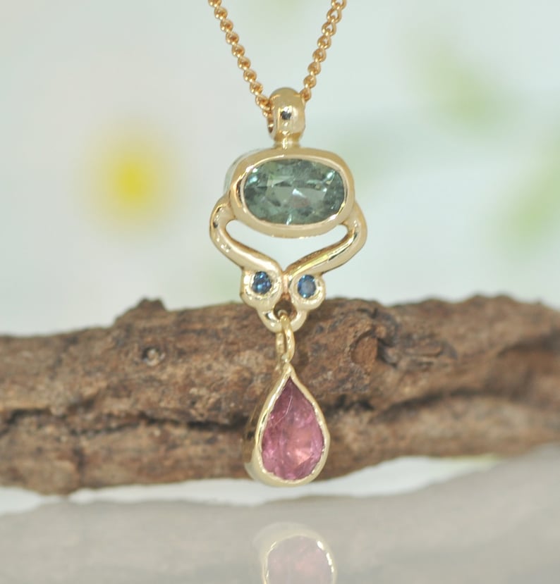 May include: A gold pendant necklace with a teardrop-shaped pink gemstone, a green oval gemstone, and two small blue gemstones. The pendant is hanging from a gold chain.