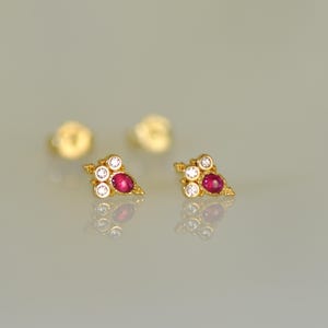 Może przedstawiać: Para złotych kolczyków sztyftów, każdy z wzorem klastra. Każdy kolczyk ma centralny owalny kamień szlachetny z rubinu, otoczony mniejszymi, okrągłymi, przezroczystymi kamieniami szlachetnymi. Kolczyki są prezentowane na odblaskowej powierzchni.