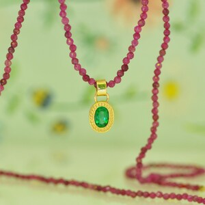 Pu&ograve; includere: Una collana con pendente in oro con una gemma verde ovale. Il pendente &egrave; sospeso a un filo di piccole perline rosa rotonde. La collana &egrave; presentata su uno sfondo morbido e sfocato.