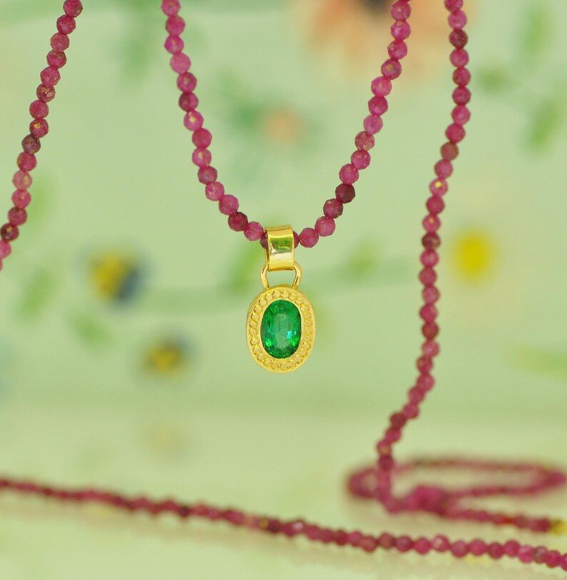 Pu&ograve; includere: Una collana con pendente in oro con una gemma verde ovale. Il pendente &egrave; sospeso a un filo di piccole perline rosa rotonde. La collana &egrave; presentata su uno sfondo morbido e sfocato.