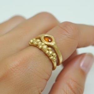 May include: Two gold rings are displayed on a finger. The top ring features an oval-shaped orange gemstone. The bottom ring has a textured design with small, round gold beads. The rings are set against a white background.