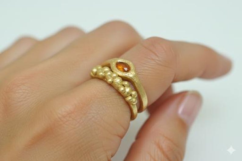May include: Two gold rings are displayed on a finger. The top ring features an oval-shaped orange gemstone. The bottom ring has a textured design with small, round gold beads. The rings are set against a white background.