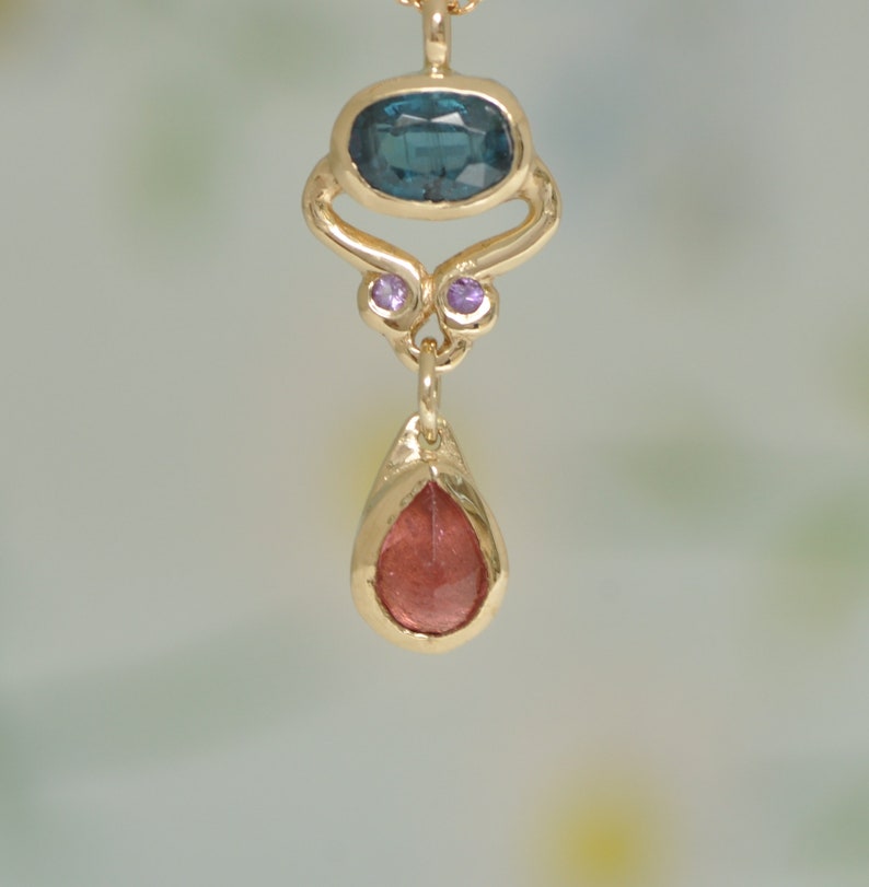 May include: A gold pendant necklace with a teardrop-shaped red gemstone, a blue gemstone, and two small purple gemstones.
