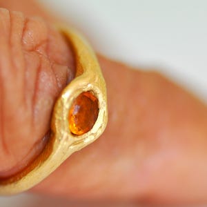 May include: A gold ring with an oval, orange gemstone. The ring has a textured, organic design. The gemstone is set in a bezel setting, and the ring appears to be handmade. The ring is on a finger.
