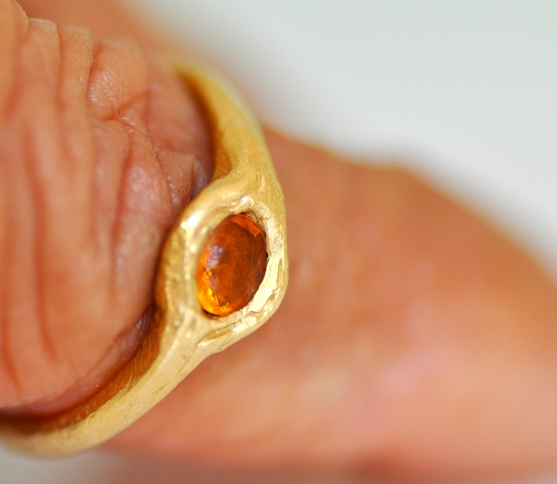 May include: A gold ring with an oval, orange gemstone. The ring has a textured, organic design. The gemstone is set in a bezel setting, and the ring appears to be handmade. The ring is on a finger.