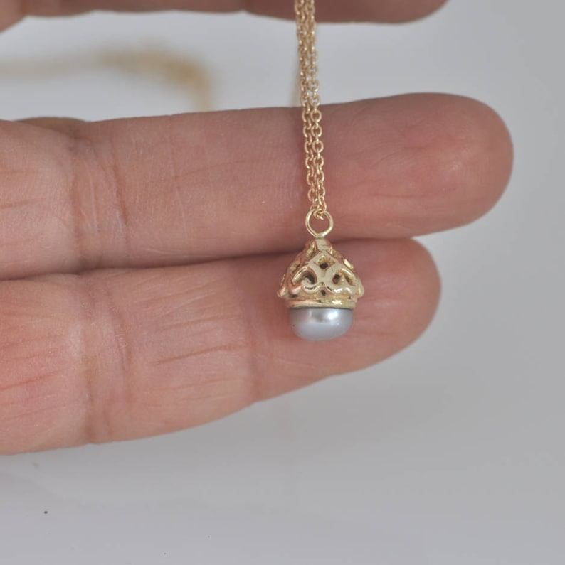 May include: A gold chain necklace with a delicate pendant featuring a gray pearl encased in a gold filigree design.