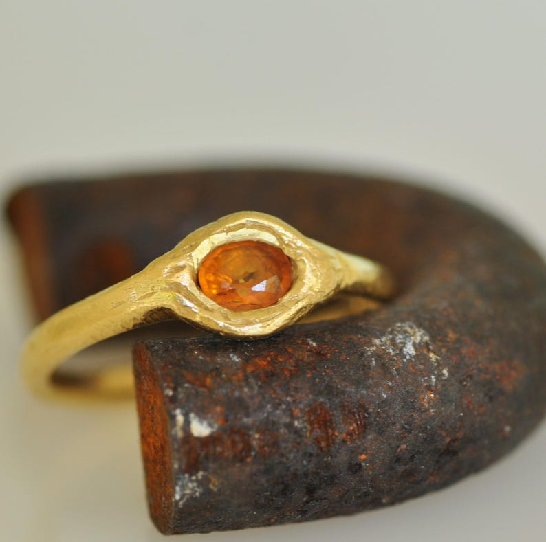 May include: A gold ring with an oval, orange gemstone. The ring has a textured band and a bezel setting that holds the gemstone. The ring is set against a dark, textured background.